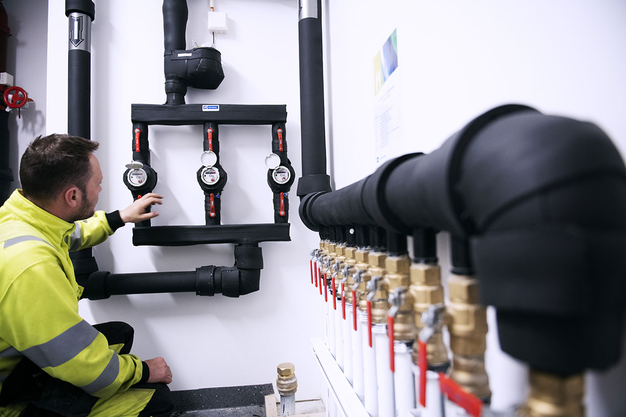 VB-rørlegger i gul refleksjakke inspiserer vannmålere i teknisk rom med svarte rør og røde kranhendler.